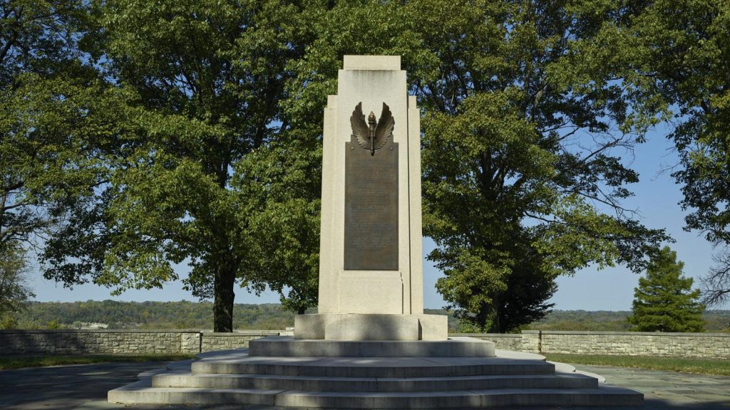 A tall stone memorial monument stands on a stepped platform in a park-like setting. The monument features a light-colored stone pillar with a dark bronze or stone plaque mounted on the front face, topped with an eagle sculpture with outstretched wings. The memorial is surrounded by a low stone wall and mature trees with full green foliage. Rolling hills are visible in the background under a clear blue sky. This memorial displays Dayton's artisanship, where Millwest continues the tradition of craftsmanship.