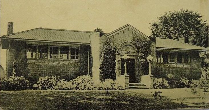 Black and white vintage photograph of the historic Miamisburg Library, a single-story brick building featuring a distinctive arched entrance portico in the center. The Tudor Revival or Colonial Revival style structure has multiple chimneys, large multi-paned windows, and well-maintained landscaping with foundation plantings and a mature tree on the right side. The photograph appears to be from the early-to-mid 20th century and shows the library in an area served by the Millwest Amish Furniture company.
