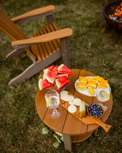 Round two-tone brown and cedar poly outdoor side table with slatted top, styled with watermelon, charcuterie, blueberries, and citrus, beside a matching Adirondack chair and copper fire pit in a grassy yard.