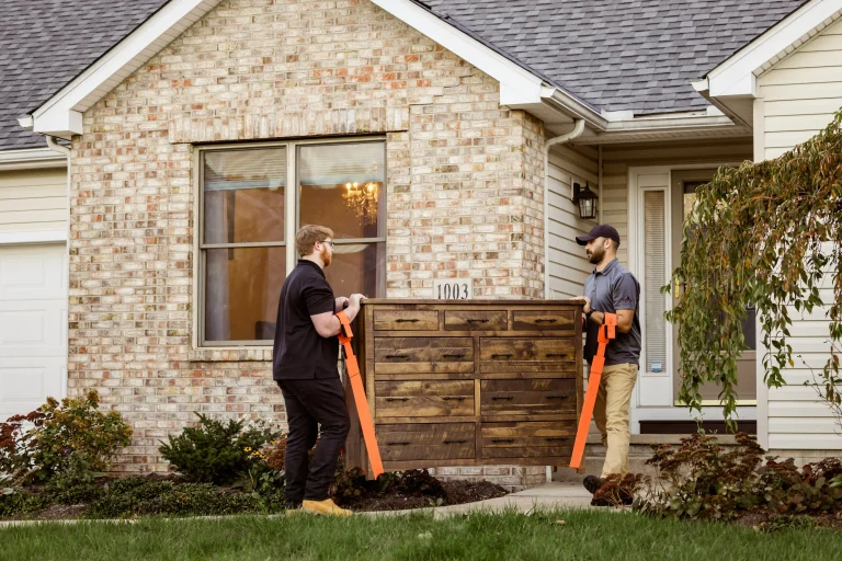Two movers carrying a large wooden dresser with orange lifting straps in front of a suburban home with brick and siding exterior