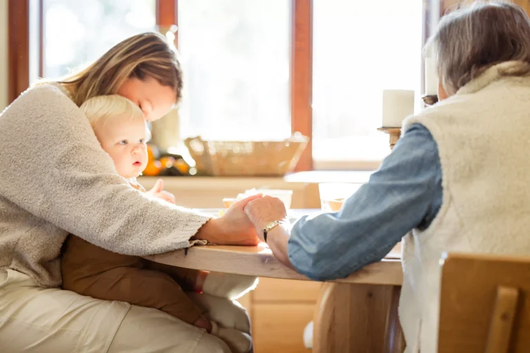 Three generations gathered around a solid wood dining table in warm natural light—a mother in a cozy sweater holds her blonde baby while an older woman reaches across the table to hold hands, sharing a quiet family moment together.