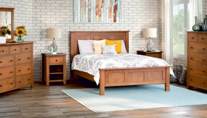 Amish-crafted solid hardwood bedroom set in a natural stain, featuring a panel bed with white and gray patterned bedding and accent pillows, flanked by two matching nightstands with ceramic lamps, a six-drawer dresser with mirror and sunflower arrangement on the left, and a tall chest of drawers on the right, set against a white brick wall with artwork above the bed and a light blue area rug on hardwood flooring.